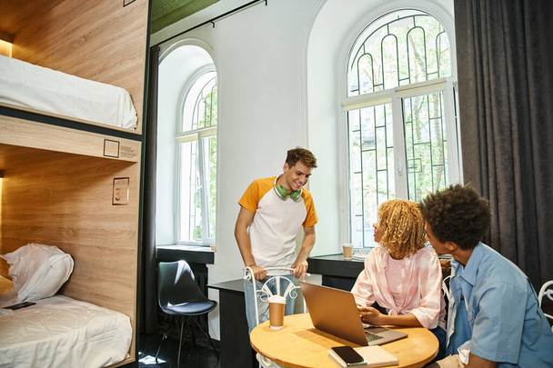 jovem estudante olhando para companheiros de quarto afro-americanos perto de laptop no aconchegante quarto albergue, viajantes - Foto, Imagem