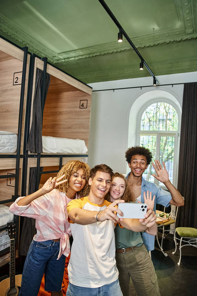 estudantes multiétnicos despreocupados acenando as mãos e tomando selfie no albergue moderno, amizade e viagens - Foto, Imagem
