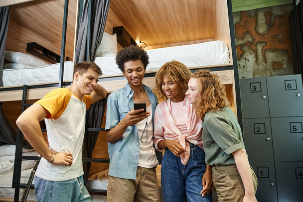glimlachende Afrikaanse Amerikaanse man browsen sociale media op smartphone in de buurt van interraciale vrienden in hostel - Foto, afbeelding