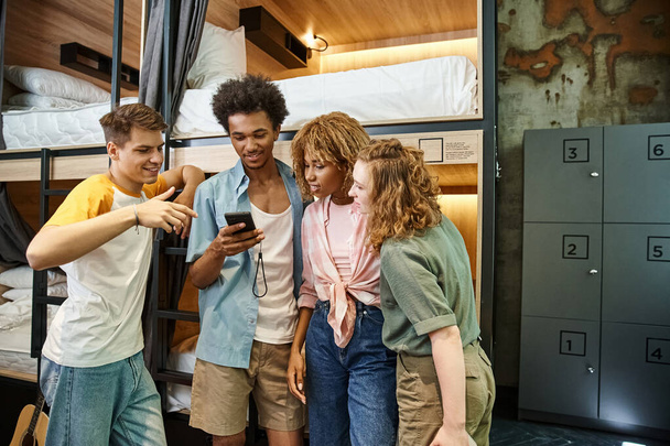 feliz estudante afro-americano usando telefone celular perto de amigos multiétnicos no albergue da juventude - Foto, Imagem
