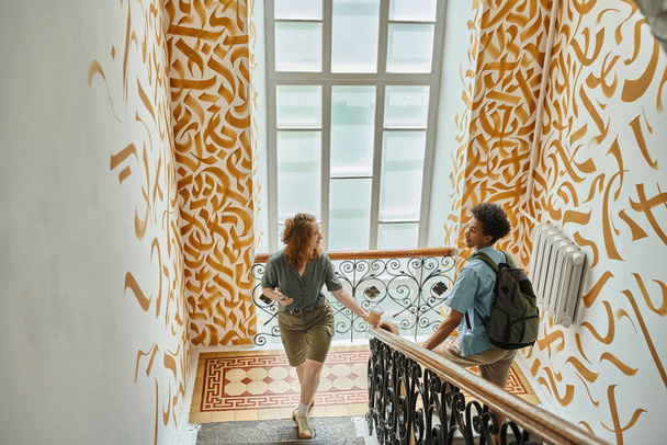 szczęśliwa kobieta i afrykański student amerykańskiego chodzenia po schodach z kolorowymi ścianami w hostelu - Zdjęcie, obraz