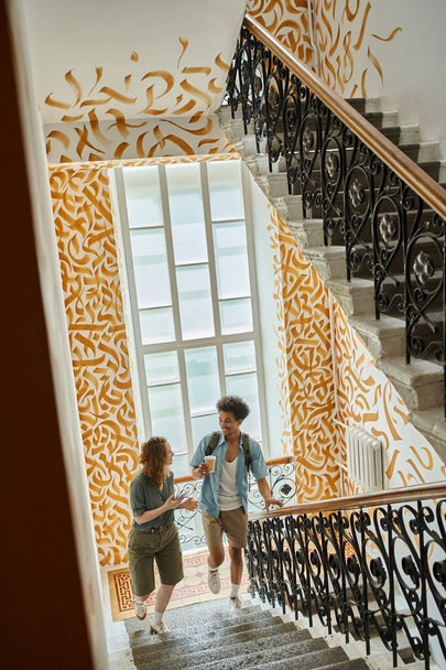 feliz pareja multiétnica caminando y hablando en la escalera con paredes de colores en albergue moderno - Foto, Imagen