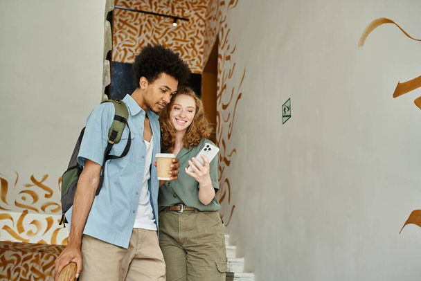 alegre pareja interracial con teléfono inteligente y café para ir caminando por las escaleras del albergue de estudiantes - Foto, Imagen