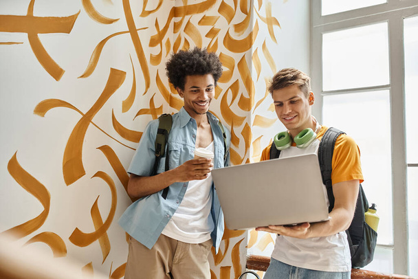 Afrikanisch-amerikanischer Student mit Pappbecher neben lächelndem Freund mit Laptop auf der Treppe des Hostels - Foto, Bild