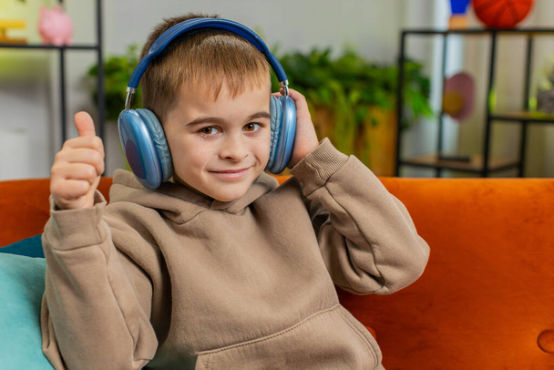 Glücklich überglückliches Teenie-Boy-Kind mit drahtlosen Kopfhörern entspannt auf der Couch im heimischen Zimmer sitzen und bevorzugte energetische Disco-Tanzmusik hören. Kinderfreizeitaktivitäten am Wochenende. Daumen hoch wie - Foto, Bild