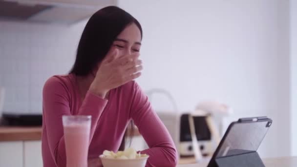 Veselý mladý asijské žena teenager v ležérní oblečení jíst nezdravé fast food snack chipsy při sledování filmu na přenosném tabletu při relaxaci na pultu kuchyně. Vysoce kvalitní 4K záběry - Záběry, video