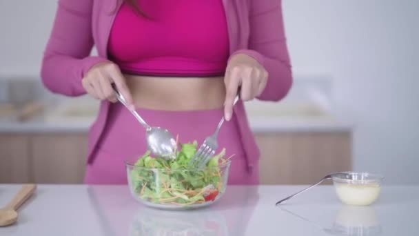 Spor giyimli genç Asyalı sporcu kadın sağlıklı bir salata yapıyor evdeki mutfakta yeşil sebzeleri karıştırarak sağlıklı bir diyet konseptini inceliyor. Yüksek kalite 4k görüntü - Video, Çekim