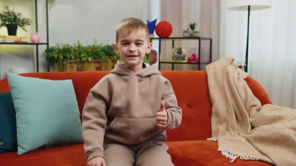 Na przykład. Szczęśliwy młody chłopiec rasy kaukaskiej patrząc z aprobatą na aparat pokazujący kciuki do góry jak znak pozytywny coś dobrego, pozytywnego sprzężenia zwrotnego. mężczyzna nastolatek mały dziecko w domu w salonie siedzi na kanapie - Materiał filmowy, wideo
