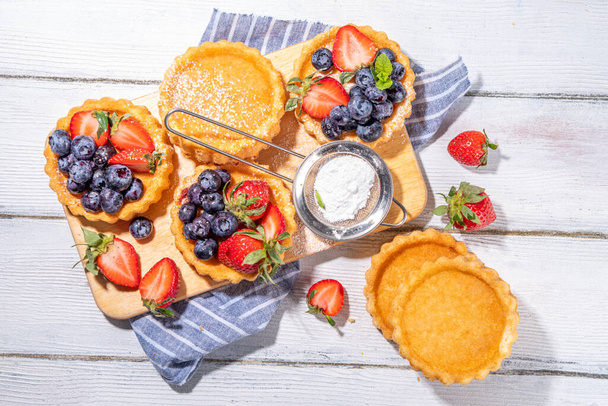 Houbové koláčky s letními bobulemi. Porcované vanilkové zákusky s čerstvými jahodami, borůvkami, malinami, na bílém dřevěném podkladovém prostoru - Fotografie, Obrázek
