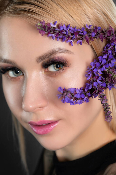 Woman's blue green eye with pupil heterochromia with beautiful make-up and thick eyebrows. Near her are beautiful blooming spring flowers. Looking at the camera. Professional makeup and cosmetology. - Photo, Image