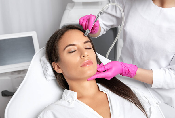 Beautiful woman with long hair, with clean fresh skin.Cosmetologist makes  procedure microdermabrasion on the face. Women's cosmetology in the beauty salon. - Photo, Image