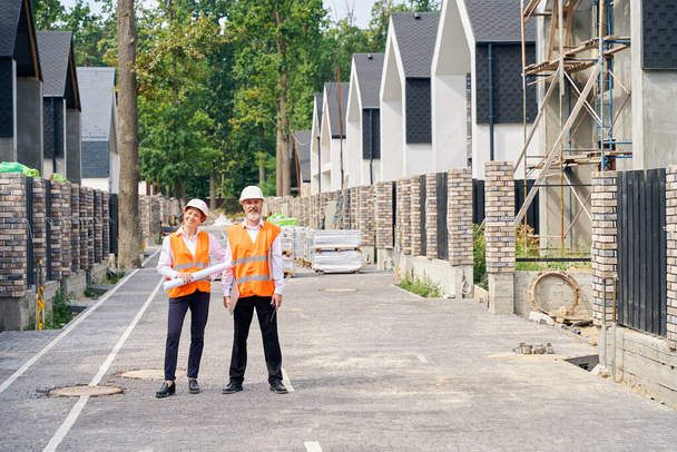 Full-length portrait of smiling engineer with blueprints and pleased foreman with walkie-talkie standing on paved street among unfinished houses - Foto, Imagem