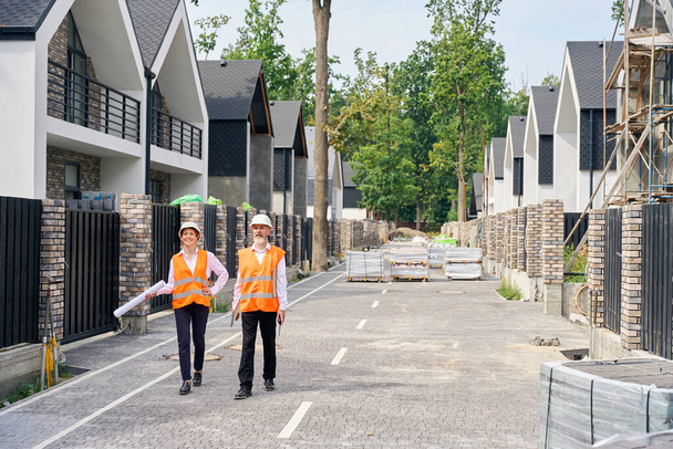 Full-size portrait of pleased female engineer and her colleague walking along paved street with unfinished private residential houses - Foto, Imagem