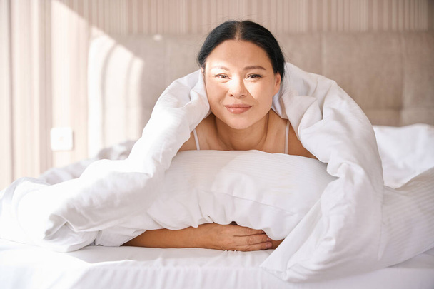 Mosolygó nő csomagolva fehér takaró feküdt kényelmes ágyban ébredés után - Fotó, kép
