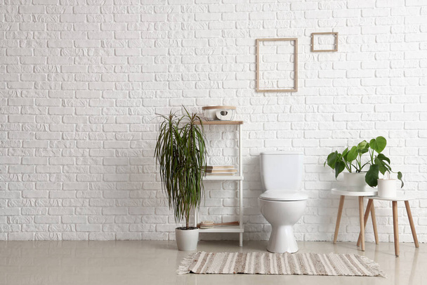 Intérieur des toilettes lumineuses avec cuvette de toilette en céramique, étagère et plantes d'intérieur près du mur de briques blanches - Photo, image