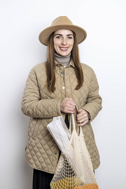 Una giovane donna in giacca con cappello tiene una borsa con generi alimentari sullo sfondo di una parete bianca - Foto, immagini