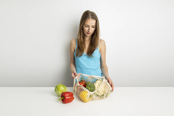 若い女性は白い背景のテーブルの上の袋から野菜を取り出します. - 写真・画像