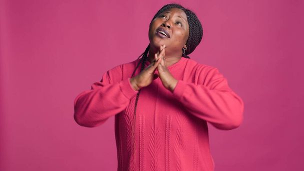 Νεαρή κυρία με αφροαμερικάνικη εθνικότητα που στέκεται και προσεύχεται με πίστη σε ροζ φόντο. Μαύρη γυναίκα σε κομψό στυλ κοιτάζοντας πάνω με τα χέρια της ψηλά δείχνοντας αφοσίωση. - Φωτογραφία, εικόνα