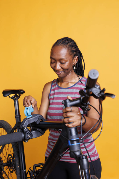 Zwarte vrouwelijke fietser inspecteert haar gebroken fietsframe terwijl ze tegen een geïsoleerde achtergrond op een reparatiestand wordt geplaatst. Jonge vrouw bereid om gespecialiseerde tools te gebruiken voor aanpassingen en reparaties. - Foto, afbeelding