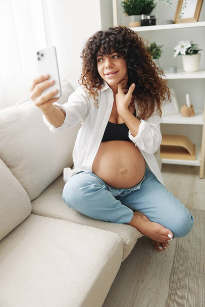 Kobieta w ciąży blogger siedzi na kanapie w domu i robi sobie zdjęcia przez telefon, selfie i rozmowy wideo, konsultacje z lekarzem online, zarządzanie ciążą. Wysokiej jakości zdjęcie - Zdjęcie, obraz