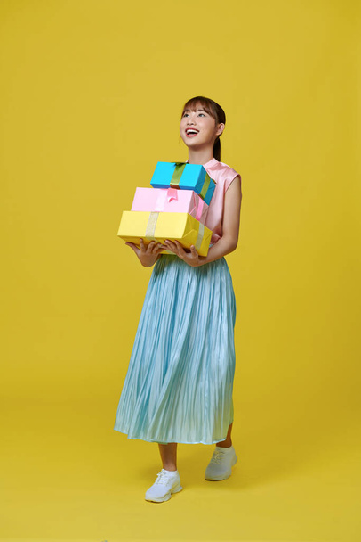 Attractive young girl in dress holding stack of gift boxes isolated over yellow background - Foto, immagini