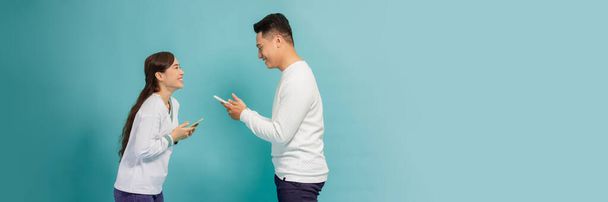 Asian girl and guy using phones sending and reading messages, standing against blue background facing each other - Photo, Image