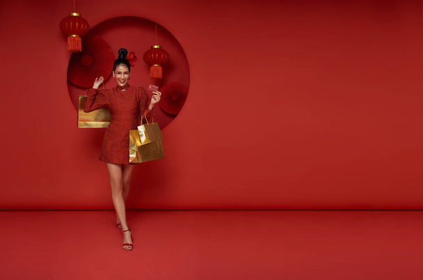 赤い背景に隔離されたショッピングバッグとクレジットカードを保持する伝統的なチョンサム qipao ドレスを身に着けている幸せなアジアのショップアホリック女性. ハッピー 中国 新年 - 写真・画像