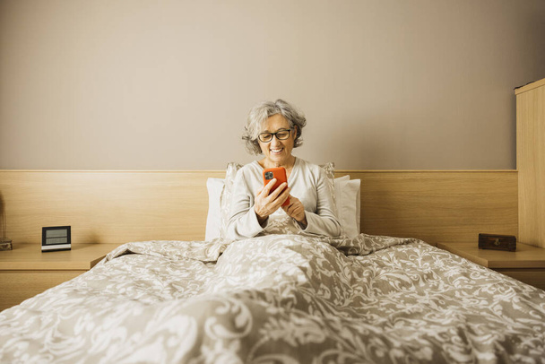 Oudere vrouw die 's morgens met een pyjama op bed zit en haar berichten bekijkt aan de telefoon.. - Foto, afbeelding