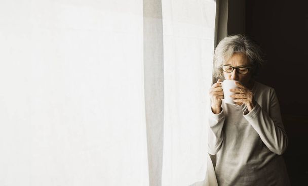 Mujer mayor parada junto a una ventana y tomando café en un día soleado en casa. - Foto, imagen