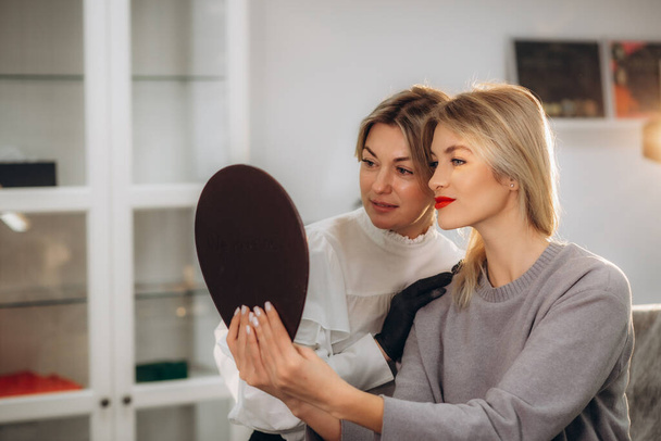 Bottom view of mirror reflection of smiling caucasian girl looking at her beautiful lips while female beautician looking at her after permanent makeup lips procedure in beauty salon. Beauty concept.  - Fotoğraf, Görsel