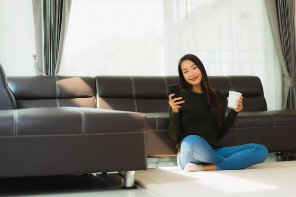 Πορτρέτο όμορφη νεαρή Ασιάτισσα γυναίκα χρησιμοποιούν έξυπνο κινητό τηλέφωνο με φλιτζάνι καφέ με καναπέ στο εσωτερικό του σαλονιού - Φωτογραφία, εικόνα