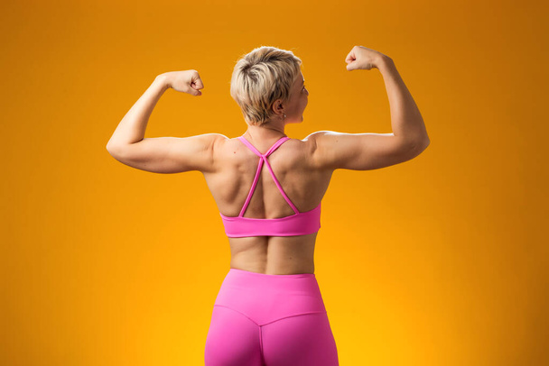 Kısa sarı saçlı, gülümseyen ve sarı arka planda kol kaslarını gösteren fitness kadını..  - Fotoğraf, Görsel