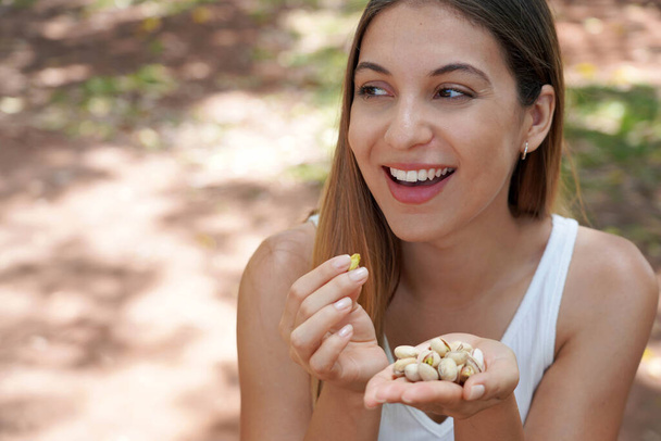 Κοντινό πλάνο μιας ελκυστικής υγιούς γυναίκας που τρώει φιστίκια στο πάρκο. Κοιτάζει στο πλάι.. - Φωτογραφία, εικόνα