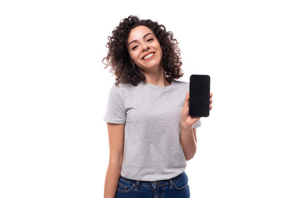 krásná radostná mladá kudrnatá bruneta žena v šedém tričku ukazuje smartphone obrazovka vertikální s mockup. - Fotografie, Obrázek