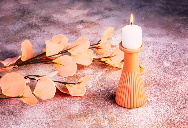 Una rama de flores artificiales en el color del año 2024 Melocotón Fuzz sobre un fondo de hormigón y un candelabro con una vela ardiente. Composición monocromática - Foto, imagen