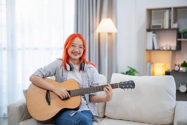 Mujer asiática con gafas y auriculares tocando la guitarra mientras se sienta en el sofá en la sala de estar en casa. Mujeres asiáticas escribiendo canciones mientras tocan la guitarra en casa. Componer concepto de música de canción. - Foto, imagen