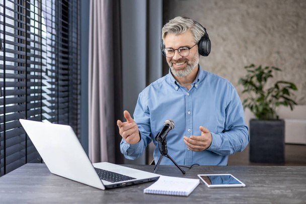 Сіроволоса старший чоловік сидить за столом вдома, носить навушники перед мікрофоном і спілкується онлайн через відеодзвінок, записуючи вебінар. - Фото, зображення
