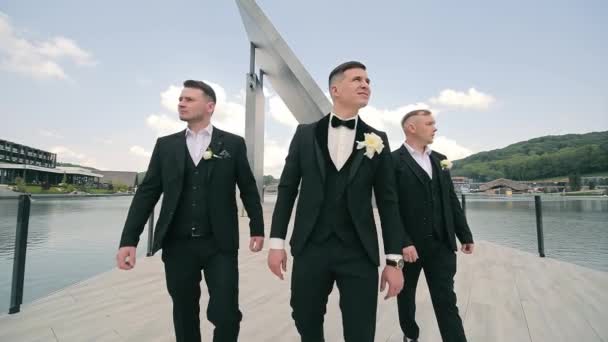Bruidsjonkers die op Dock lopen. Drie bruidsjonkers in zwarte pakken lopen op een steiger bij het meer. - Video