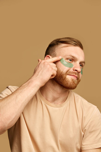Close up portrait of young attractive man applies hydrogel patches under the eyes against swelling and bruising against beige background. Concept of beauty treatment, selfcare and hygiene, cosmetic. - Foto, immagini