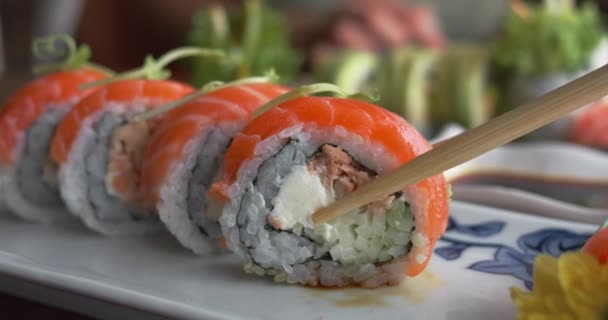 Jím sushi v restauraci s hůlkami. Zblízka hůlky uchopit červené sushi rohlíky, namočené v sójové omáčce jíst s gustem a požitkem. Krásné vkusné sushi zdobené zelení jedl - Záběry, video