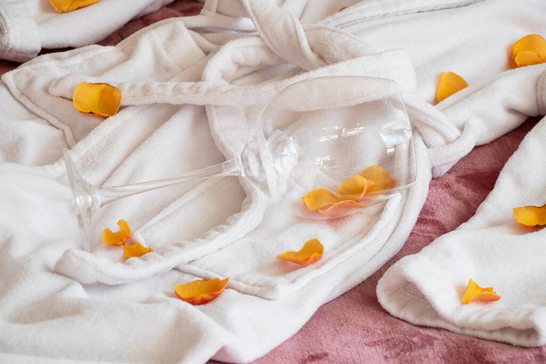 Жіночий білий махровий халат лежить на ліжку, розкидані пелюстки троянд і порожній келих вина, романтичні відносини - Фото, зображення