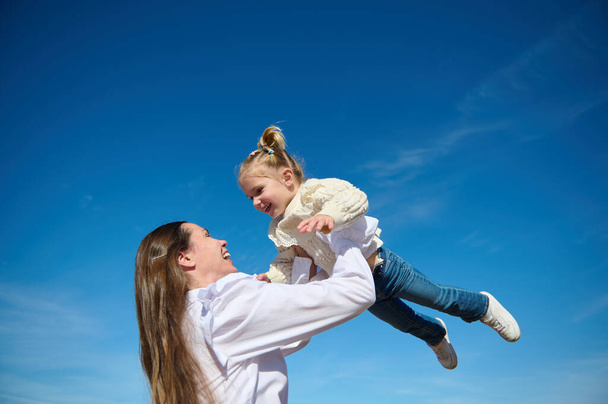 Κάτω όψη μιας ευτυχισμένης αρμονικής οικογένειας μιας νεαρής στοργικής μητέρας που ξερνάει το μωράκι της, ένα χαριτωμένο κοριτσάκι, γελώντας και παίζοντας το καλοκαίρι στη φύση με φόντο τον γαλάζιο ουρανό - Φωτογραφία, εικόνα