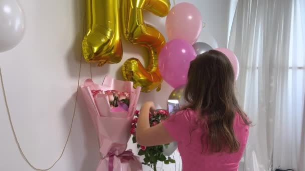 A lány képeket készít az ajándékairól, virágokról és lufikról a telefonján. ünneplik 15. születésnapját, tinédzser lány szaladgál a szobában húzza hélium lufik szelfi - Felvétel, videó