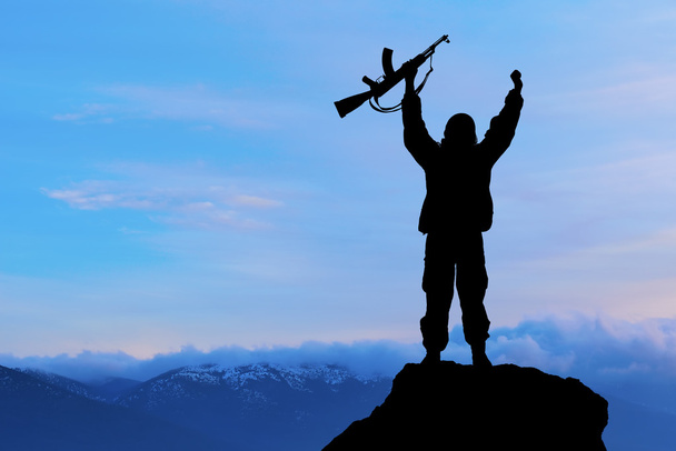 Sziluettjét katonai katona vagy tisztviselő fegyverekkel, naplementekor. lövés, holding a fegyvert, színes égbolt, a hegyi háttér - Fotó, kép