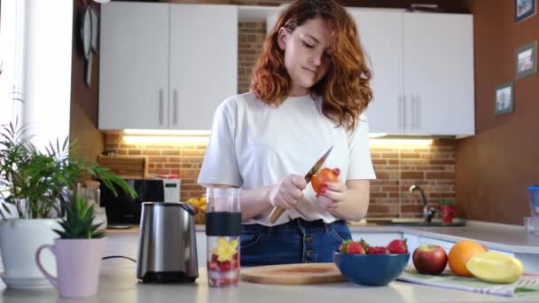 Młoda kobieta w domu w kuchni zrobić koktajl owocowy. Piękna młoda, szczęśliwa kobieta w domowej kuchni przygotowuje koktajl fitness w domu. Mączka sportowa i wegańska - Materiał filmowy, wideo