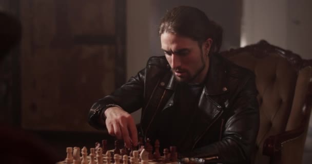Серьезный взрослый мужчина в кожаной куртке сидит на стуле и играет дома в шахматы - Кадры, видео