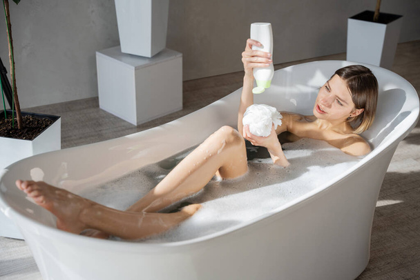 Junge blonde Frau gießt Körperwäsche auf Schwamm, während sie sich in der Badewanne ausruht. Blick von oben auf eine entspannte Frau mit einer Flasche Duschgel, während sie zu Hause badet. Hygiene, Kosmetikkonzept. - Foto, Bild