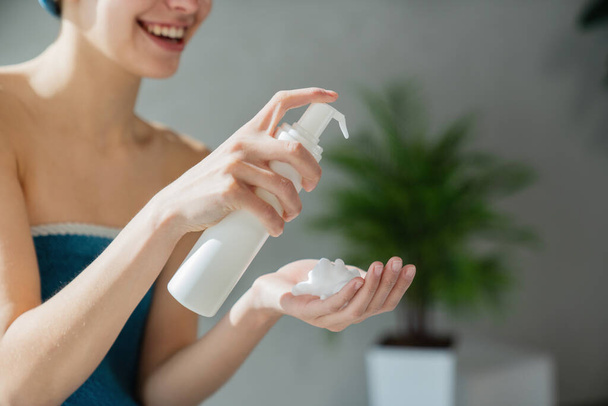 Boldog nő alkalmazása habos szappan fehér műanyag palack a fürdőszobában. Közelkép egy névtelen lányról, aki habos folyadékot ad a tenyerébe, homályos háttérrel. A bőrápolás fogalma. - Fotó, kép