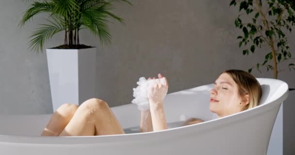Спокійна красива жінка миє губкою, одночасно замочуючи у стильній ванні. Вид збоку розслабленої блондинки, яка стискає губку, приймаючи ванну в розкішному готелі, в повільному русі. Розслабтеся і спа концепція. - Кадри, відео