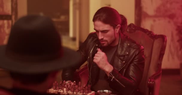 Real time handheld shot of thoughtful young male Mexican with ponytail looking away while sitting at table with unrecognizable opponent guy in hat and moving black pawn on chessboard - Footage, Video